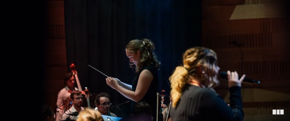 A conductor holds a baton and score while leading an orchestra on stage, with a singer at a microphone in the foreground.