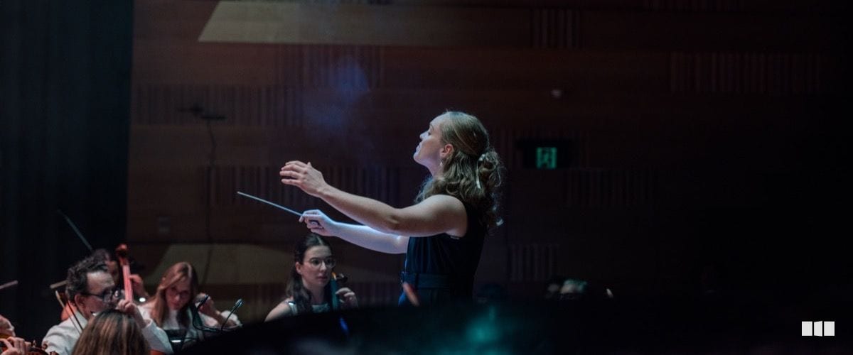 A female conductor leads an orchestra with expressive gestures and baton raised during a concert performance.