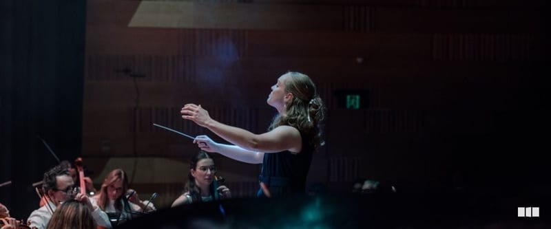 A female conductor leads an orchestra with expressive gestures and baton raised during a concert performance.