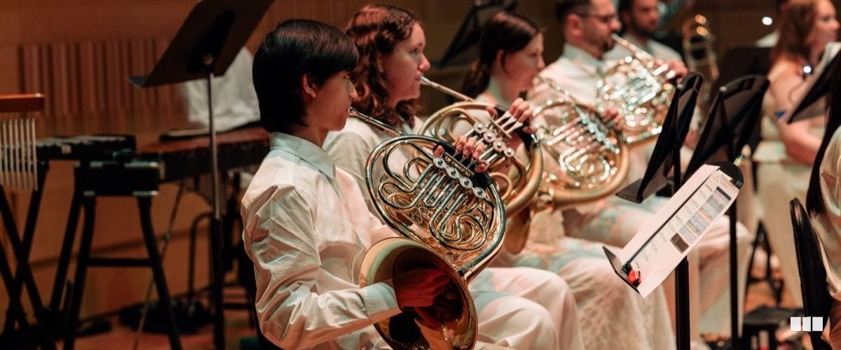 A section of French horn players in white attire perform together during an orchestral concert, reading from music stands.