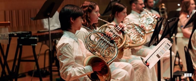 A section of French horn players in white attire perform together during an orchestral concert, reading from music stands.
