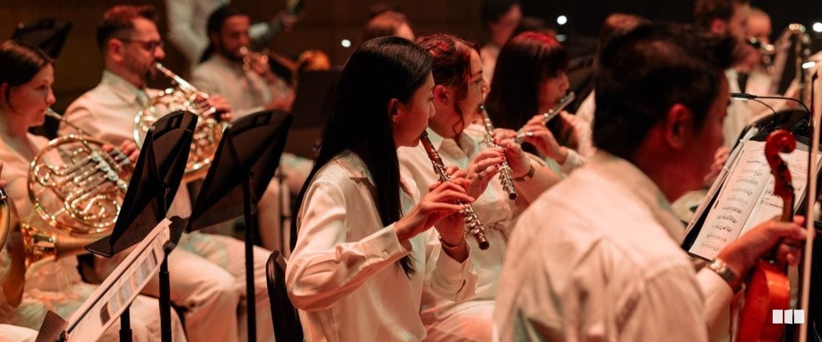 Orchestra musicians in white attire perform woodwind and brass instruments during a concert, reading from music stands.