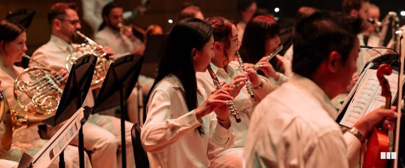 Orchestra musicians in white attire perform woodwind and brass instruments during a concert, reading from music stands.