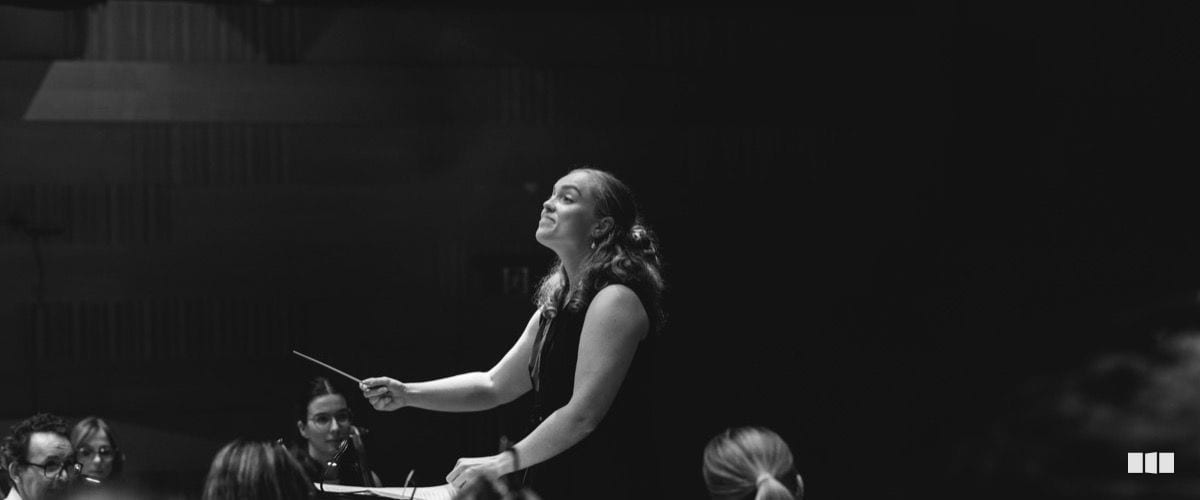 A female conductor leads an orchestra with expressive energy, baton raised, during a performance in a concert hall.