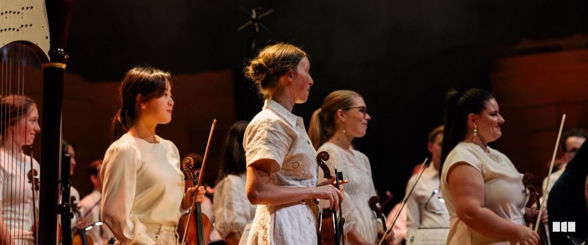 Female string players in white dresses stand holding violins during an orchestral bow on a warmly lit concert stage.