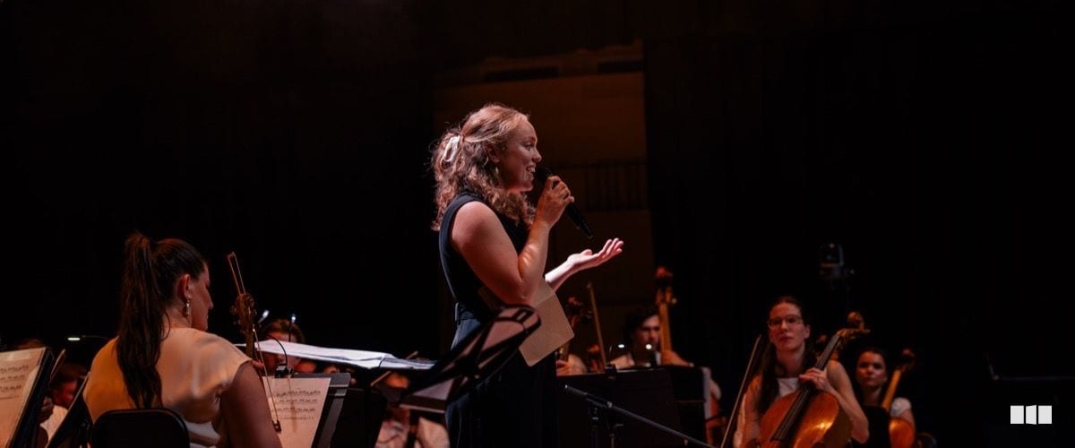 A female soloist sings into a handheld microphone on stage while orchestra musicians play behind her in warm stage lighting