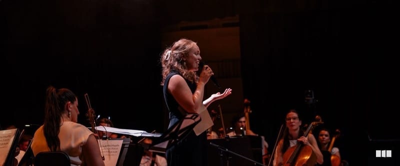 A female soloist sings into a handheld microphone on stage while orchestra musicians play behind her in warm stage lighting