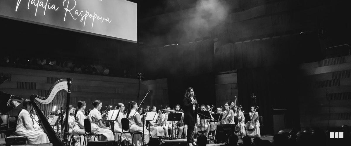 Soloist Natalia Raspova performs with a full orchestra on a darkened concert stage, with her name displayed on a large screen behind the ensemble