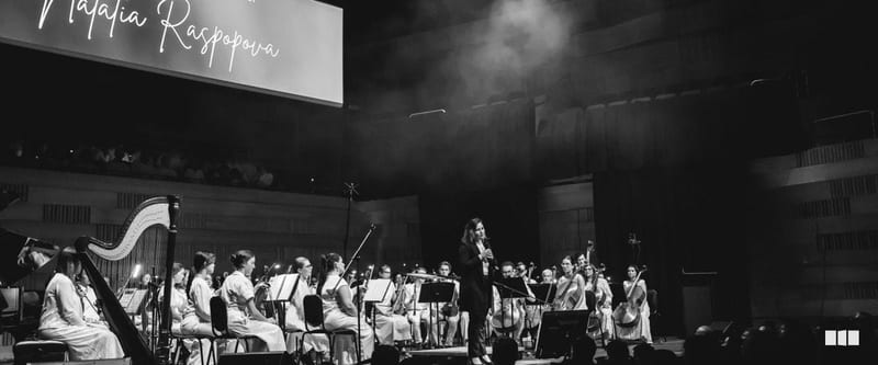 Soloist Natalia Raspova performs with a full orchestra on a darkened concert stage, with her name displayed on a large screen behind the ensemble
