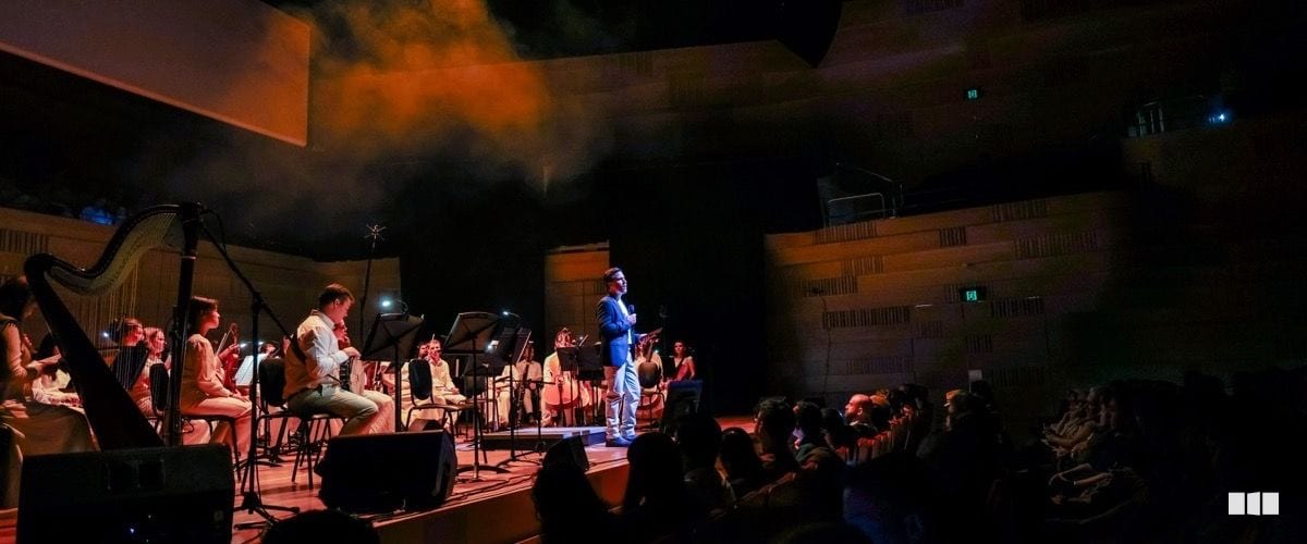A conductor in a blue jacket addresses an orchestra on a warmly lit concert stage with dramatic orange smoke effects above