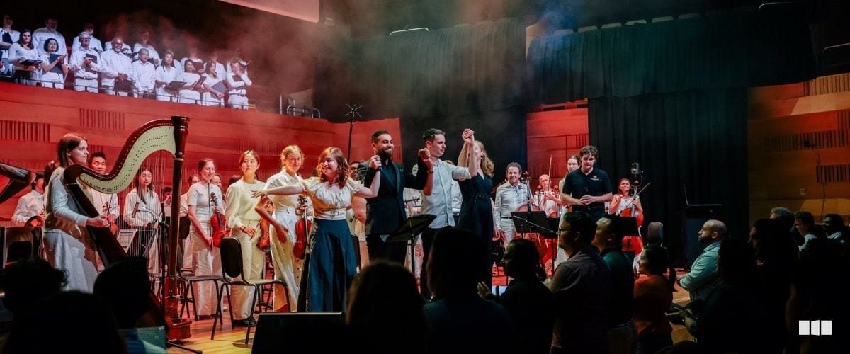Orchestra and choir take a celebratory bow together on a red-lit concert stage after a performance, with soloists raising joined hands at centre stage