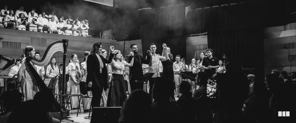 Orchestra and choir members take a bow together on stage at the conclusion of a concert, with several performers raising joined hands in celebration