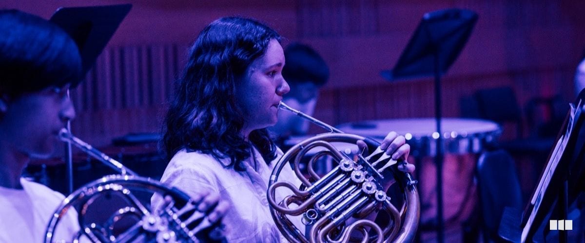 A young female musician plays French horn in a youth orchestra rehearsal under atmospheric blue-purple stage lighting.