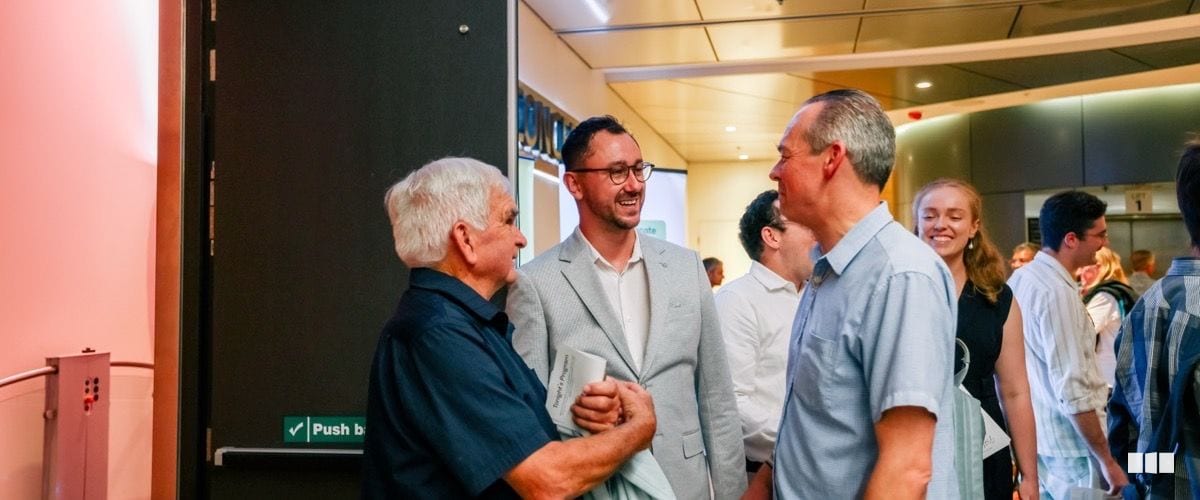 Three men share a laugh in conversation during a post-concert foyer gathering, with other attendees visible in the background