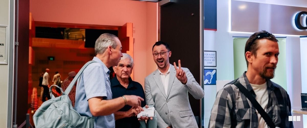Four men chat and laugh in the foyer of a performance venue near an audio assistance sign