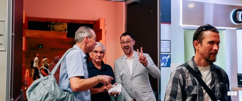 Four men chat and laugh in the foyer of a performance venue near an audio assistance sign
