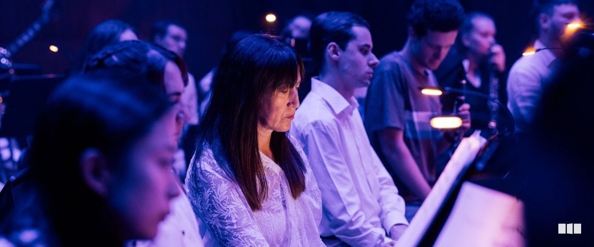 A woman with dark hair and a white lace blouse sits at the piano alongside other musicians during a dimly lit orchestral performance