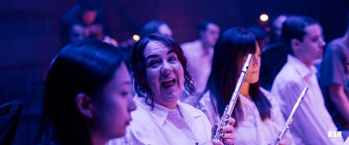 A flautist laughs expressively mid-rehearsal while fellow musicians holding flutes sit around her in purple-lit concert hall.