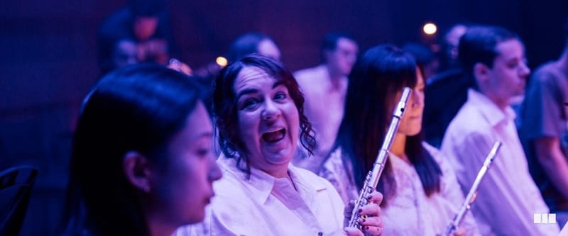 A flautist laughs expressively mid-rehearsal while fellow musicians holding flutes sit around her in purple-lit concert hall.