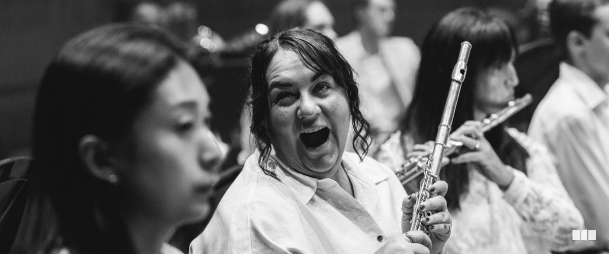 A flautist laughs expressively mid-rehearsal while holding her flute, surrounded by fellow orchestra musicians