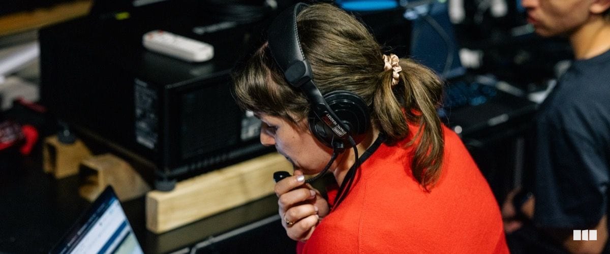 A woman wearing headphones and a red top speaks into a handheld radio or intercom device at a technical operations station