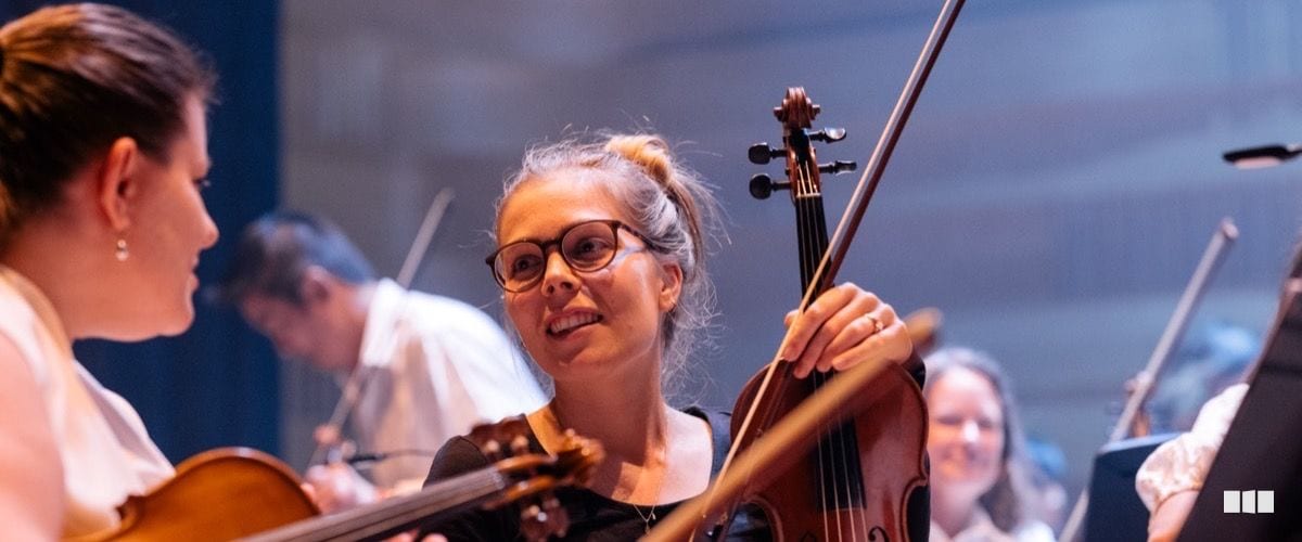A smiling violinist wearing glasses looks up during an orchestra rehearsal while holding her instrument and bow