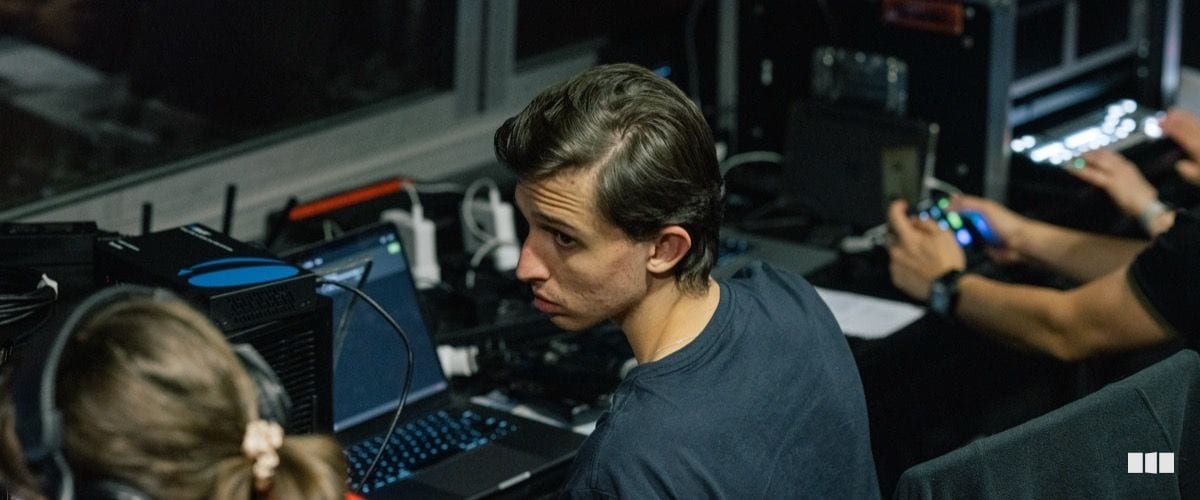 A young audio or lighting technician focuses intently on a laptop at a technical control station during a performance or rehearsal.