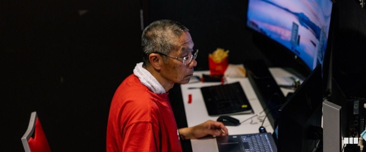An older man wearing a red shirt and glasses works intently at a computer workstation with a large monitor in a darkened room