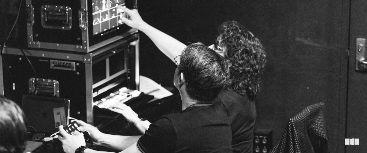 Two production crew members operate a video switcher and mixing console backstage during a live event.