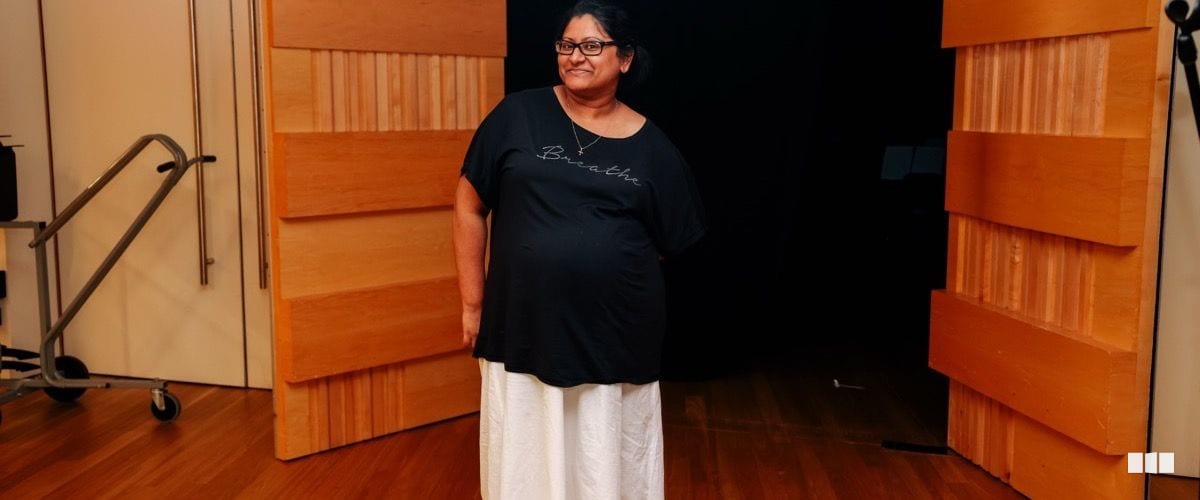A smiling woman wearing a black 'Breathe' t-shirt and white skirt stands in front of wooden acoustic panels on a stage
