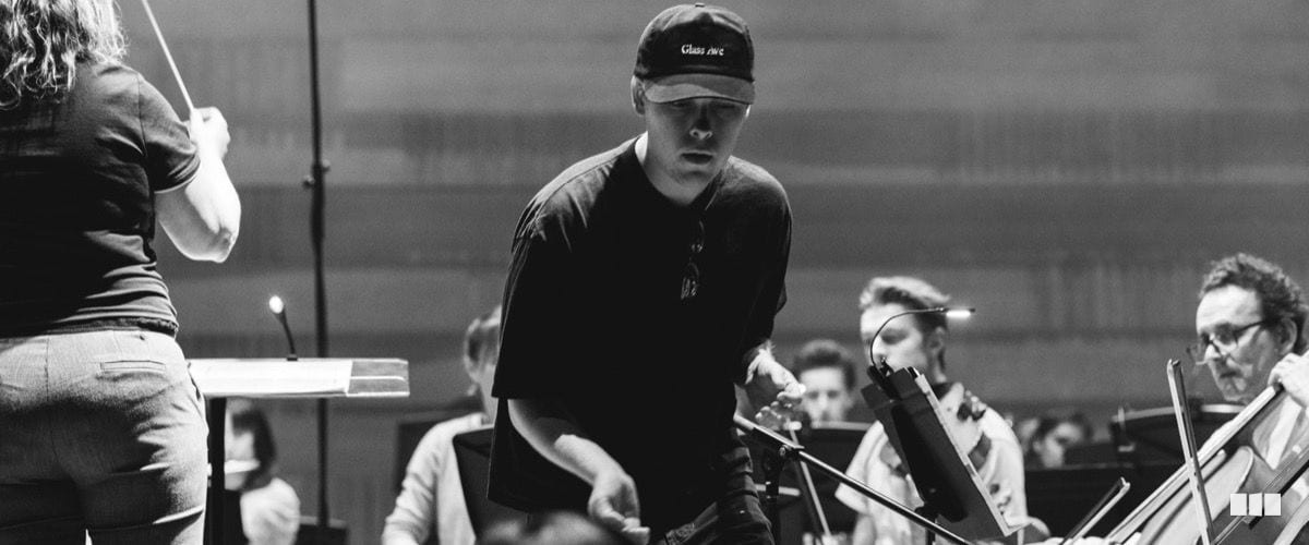 A person in a black cap and t-shirt leans over a music stand during an orchestra rehearsal while a conductor raises a baton in the foreground.