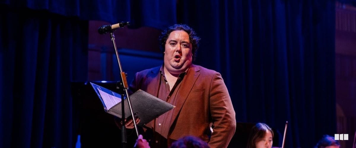 A male soloist in a brown blazer sings into a microphone while holding a black score folder on a darkly lit stage, with orchestral musicians visible in the background.