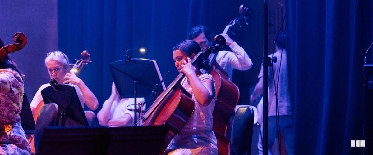 A young cellist performs alongside other string players in a blue-lit concert setting, reading from music stands.