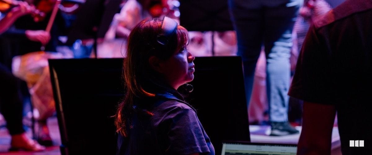 A woman wearing a headset and VR-style glasses looks upward attentively while seated among orchestra musicians on stage under dramatic purple and red lighting