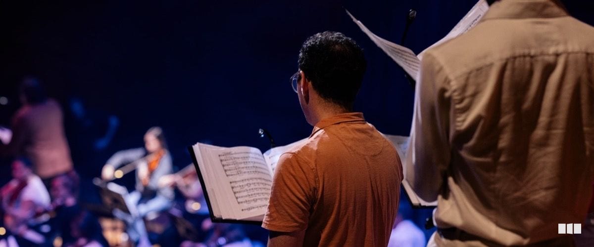 Two musicians stand with open sheet music on stands, viewed from behind, as the orchestra rehearses on a darkened stage.