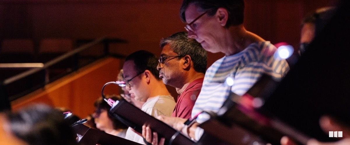 Choir members follow their scores under music stand lights during a rehearsal in a concert hall