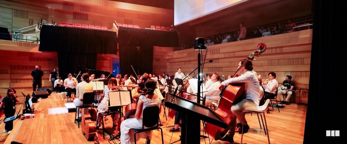 An orchestra ensemble rehearses or performs on a warm timber-panelled concert hall stage, viewed from behind the musicians with an audience visible in the upper gallery