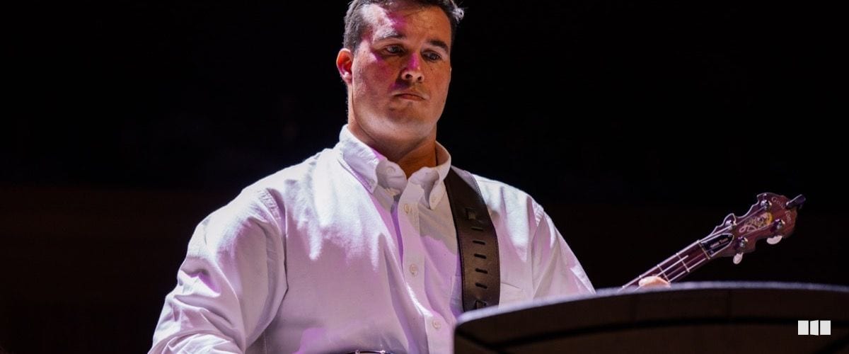 A young male musician in a white button-up shirt stands at a podium with a guitar strapped across his shoulder, lit by warm stage lighting against a dark background.