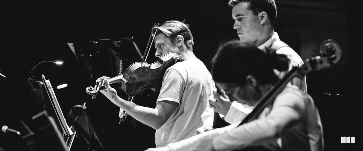 Three string musicians perform together in focused concentration during an orchestral rehearsal or concert, reading from music stands.