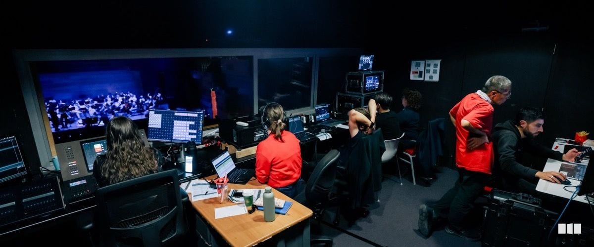 Production crew members work at technical consoles in a control booth overlooking an orchestra performing on stage