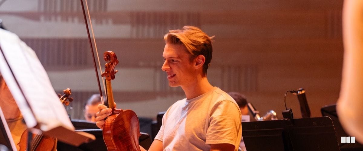 A young male violinist smiles warmly while holding his violin and bow during an orchestral rehearsal