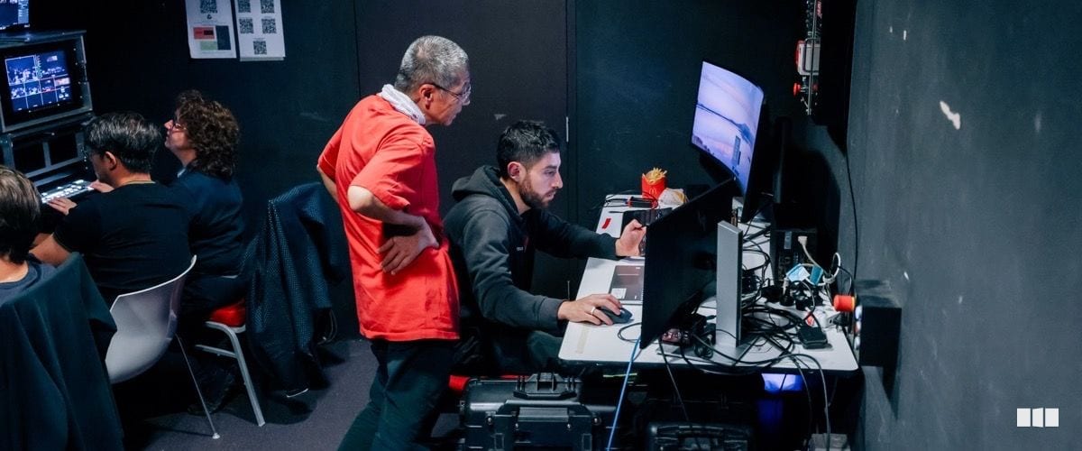 Two crew members review footage or technical content on a monitor at a production control desk backstage, with additional crew visible working at equipment in the background.