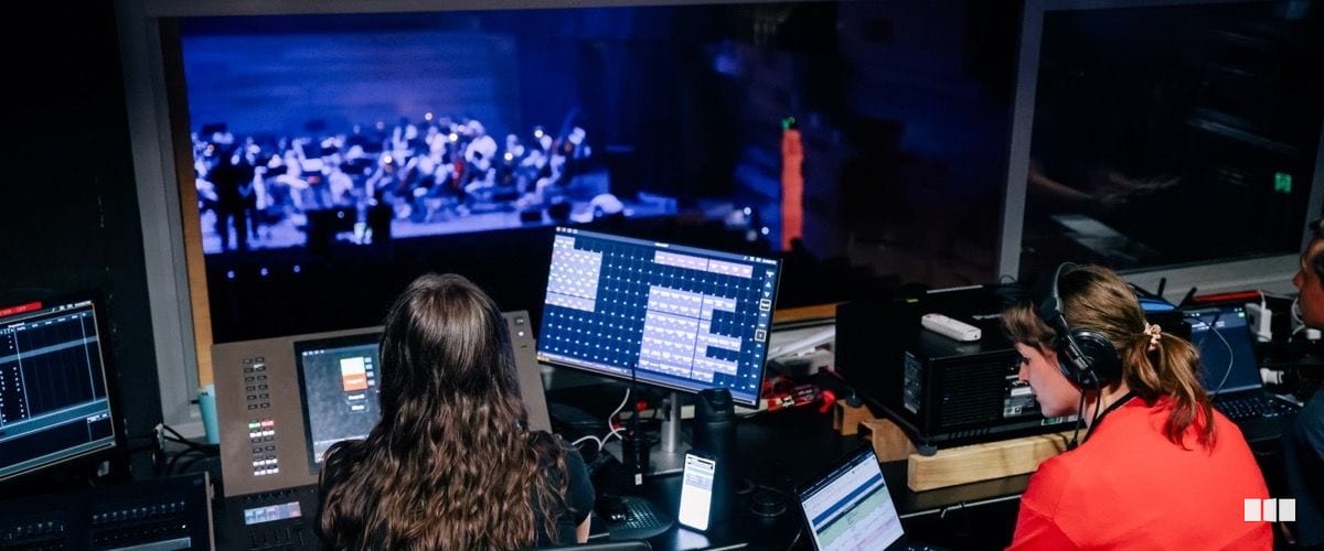 Two production crew members operate lighting and audio consoles in a technical booth overlooking an orchestra rehearsal on stage.