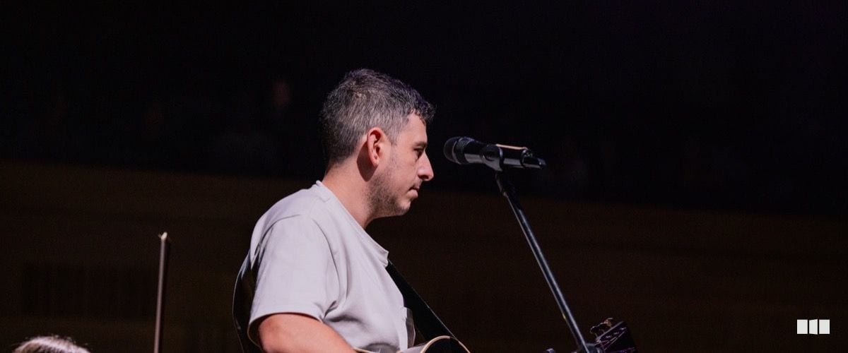 A man with greying hair plays acoustic guitar and sings into a microphone on a darkened stage