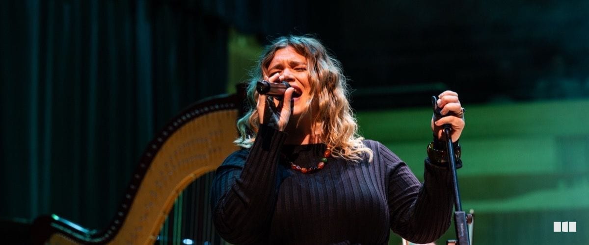 A female vocalist performs passionately on stage with a harp visible in the background under dramatic stage lighting.