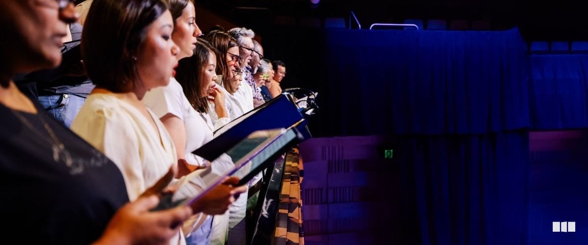 A row of choir singers stand in profile holding blue folders and singing during a performance on a darkened stage.