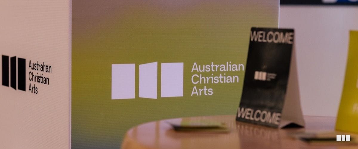 Australian Christian Arts branding displayed on a curved exhibition backdrop alongside a black welcome tent card on a table