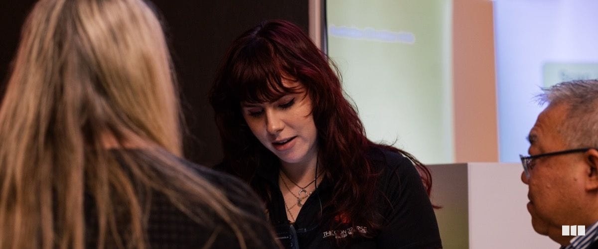 A young woman with dark red hair speaks to attendees during what appears to be a presentation or workshop setting