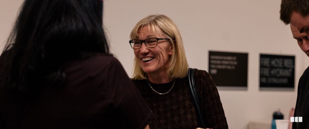 A smiling blonde woman wearing glasses and a dark patterned top engages warmly in conversation at what appears to be a post-concert reception or gallery event