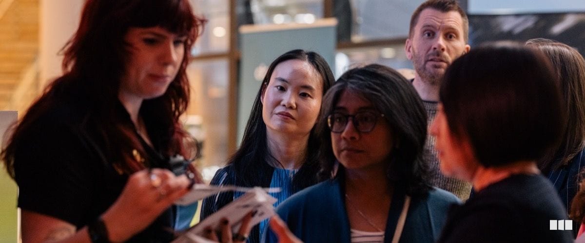 A small group of people gathered closely together in conversation, with one woman appearing to demonstrate or show something in her hands at what looks like a professional event or exhibition.
